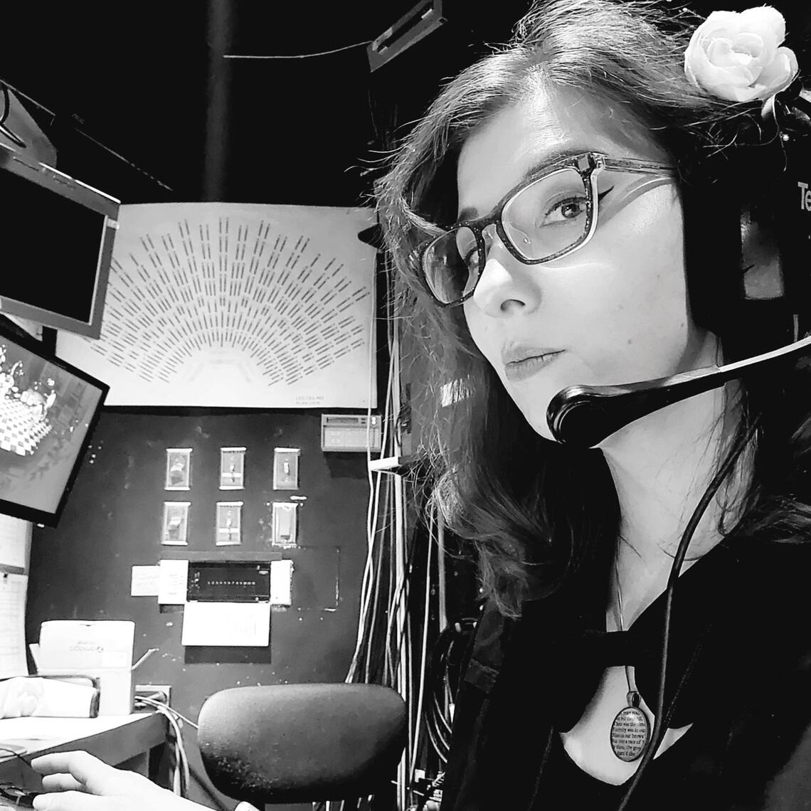 Mackenzie in a booth calling a show. She wears a large headset and a flower in her hair.